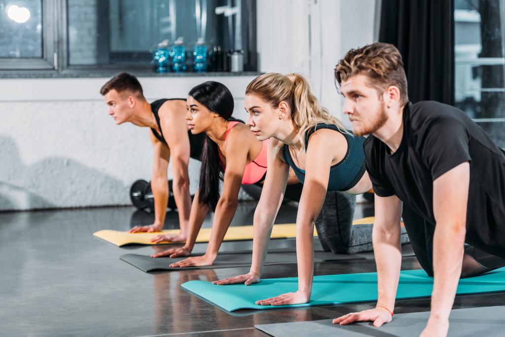 hombres y mujeres jóvenes atléticos multiétnicos haciendo ejercicio sobre esterillas de yoga y mirando hacia otro lado en el gimnasio
 - Foto, Imagen