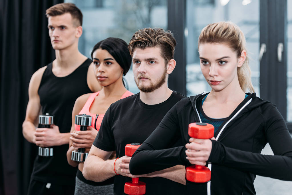 multiethnic sporty young friends holding dumbbells and looking away in gym - Photo, Image