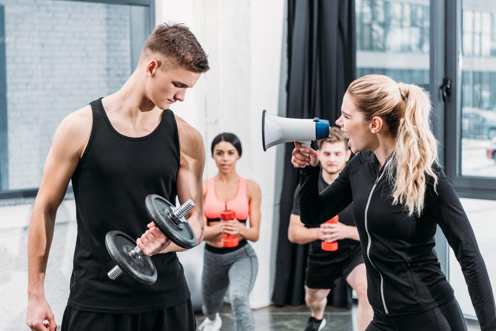sportswoman with megaphone yelling at young man training with dumbbell in gym - Photo, Image