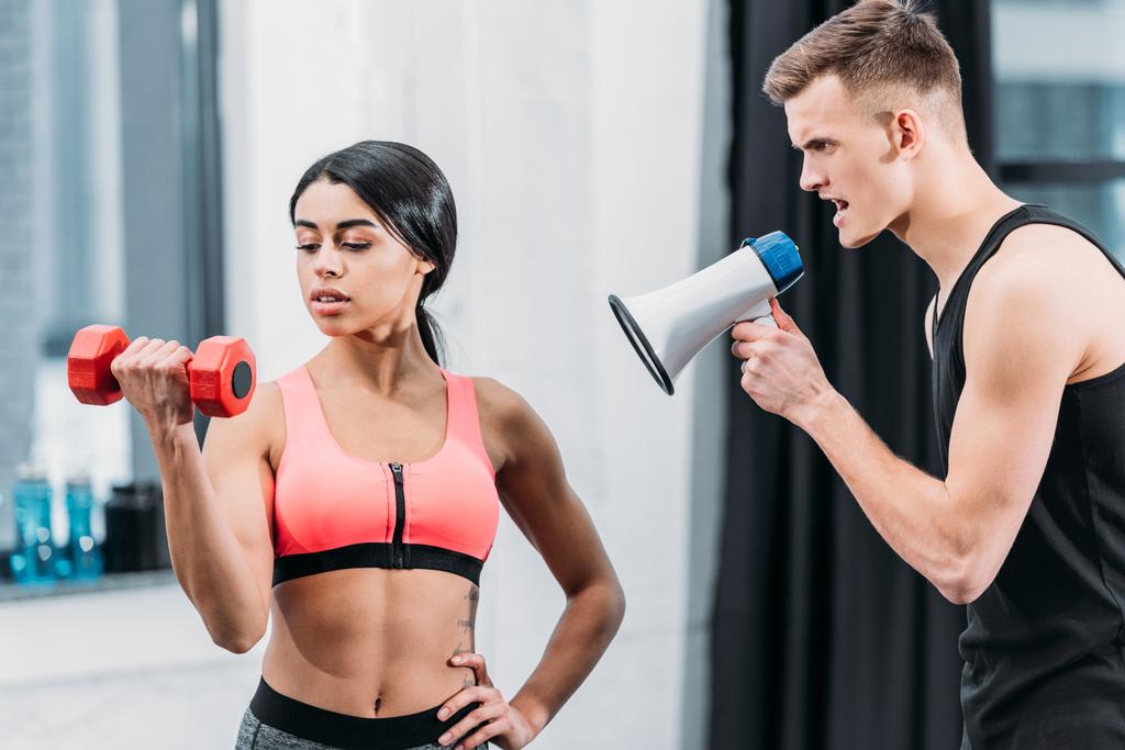 trainer with megaphone yelling at sporty african american girl training with dumbbell in gym - Photo, Image