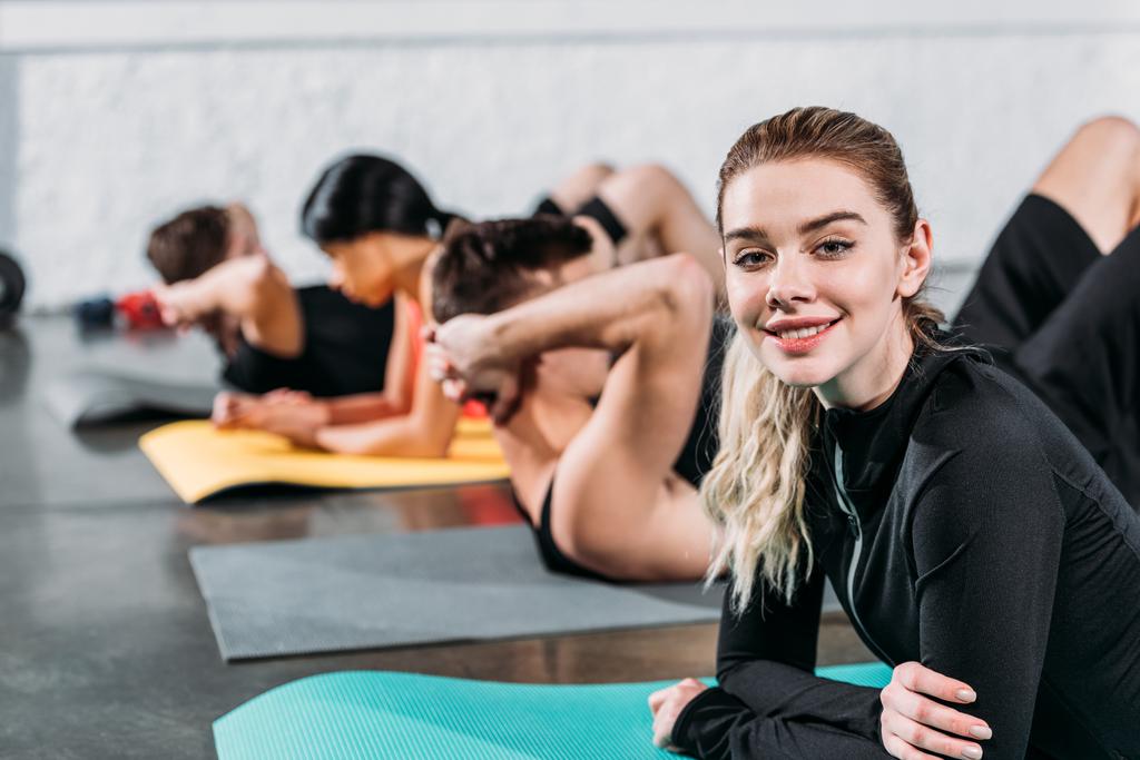 krásná mladá žena leží v jóga mat a usmívá se na kameru při tréninku s přáteli v tělocvičně - Fotografie, Obrázek