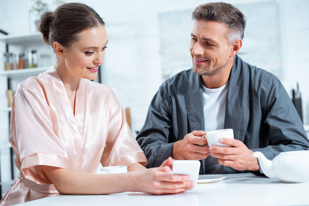 piękna para uśmiechający się w szaty przy użyciu smartfona mając herbaty podczas śniadania w kuchni - Zdjęcie, obraz