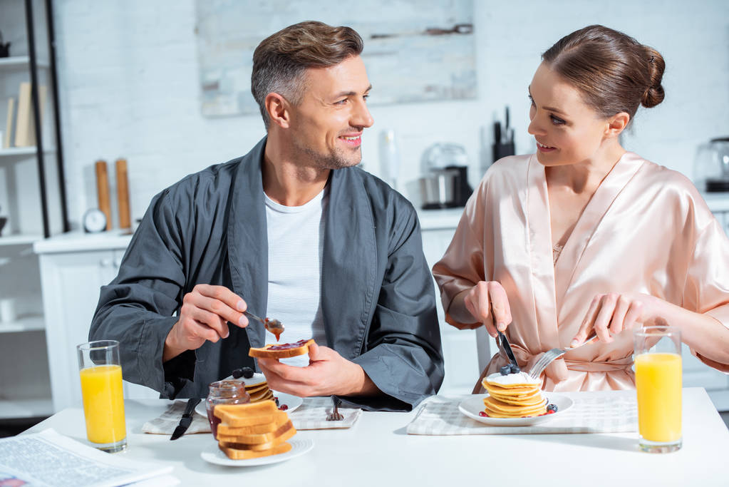 Lächelndes erwachsenes Paar in Bademänteln beim Frühstück mit Pfannkuchen und Orangensaft in der Küche - Foto, Bild