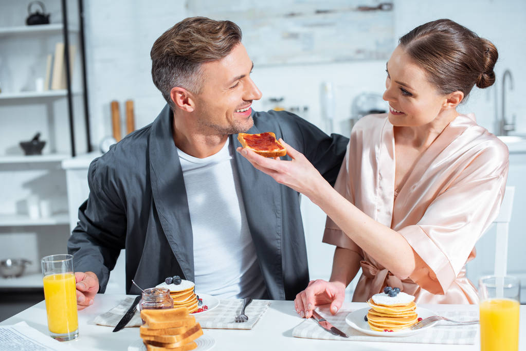 volwassen paar in gewaden te glimlachen tijdens het ontbijt met pannenkoeken en sinaasappelsap in keuken - Foto, afbeelding