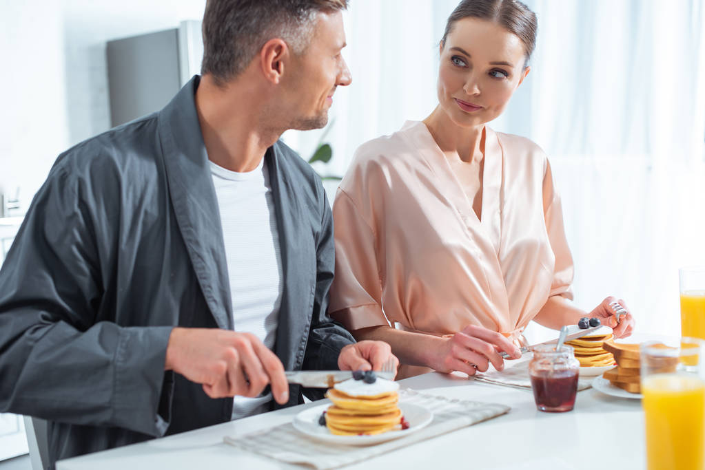 hermosa pareja en túnicas durante el desayuno con panqueques y jugo de naranja en la cocina
 - Foto, Imagen