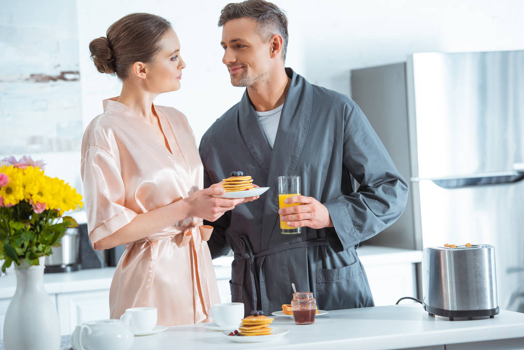 schönes Paar in Bademänteln mit Orangensaft und Pfannkuchen beim Frühstück in der Küche - Foto, Bild