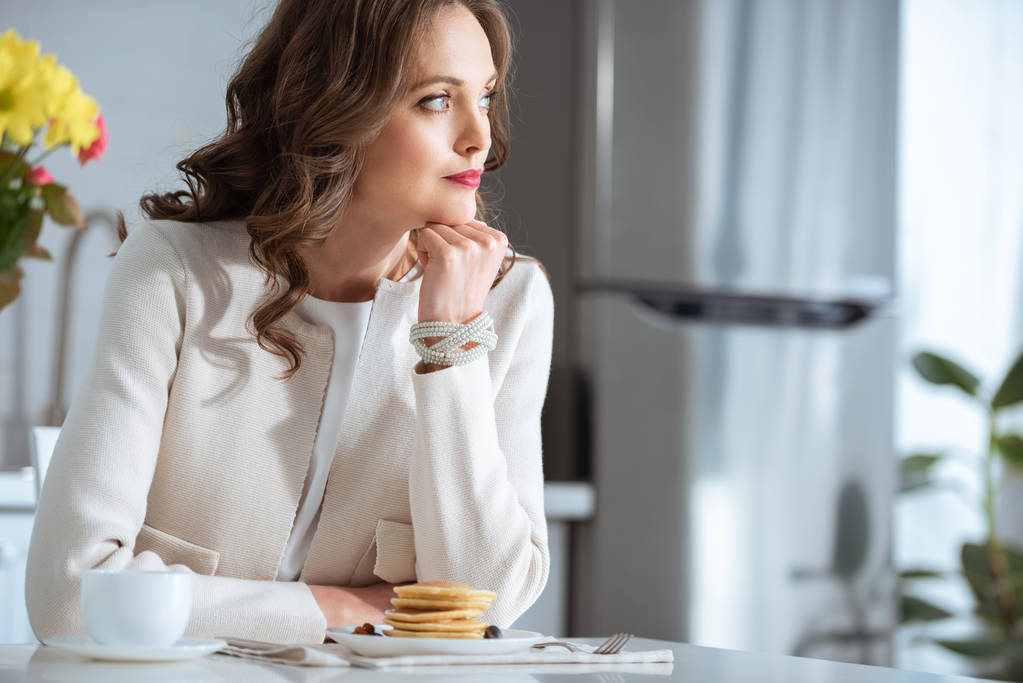 όμορφη γυναίκα συλλογισμένος κάθεται στο τραπέζι με τηγανίτες και καφέ κατά τη διάρκεια του πρωινού στην κουζίνα - Φωτογραφία, εικόνα