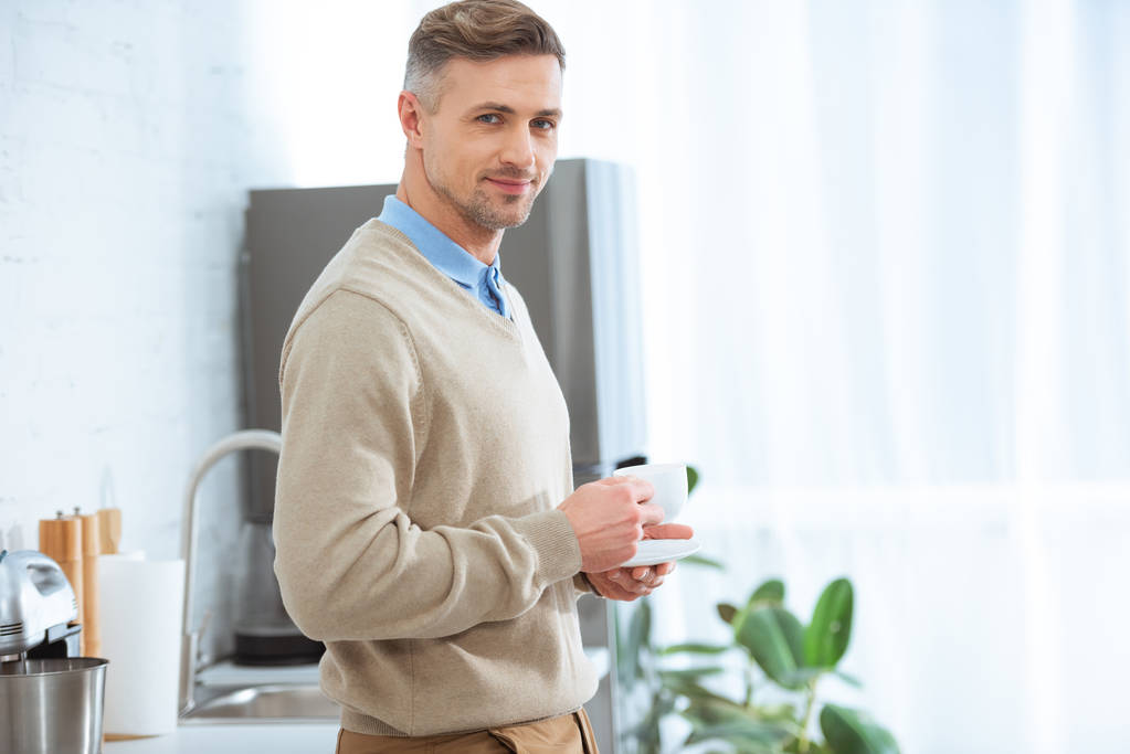 pohledný muž v neformálním oblečení drží šálek kávy a při pohledu na fotoaparát v kuchyni - Fotografie, Obrázek