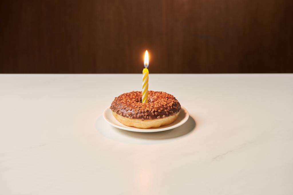 burning candle in middle of sweet doughnut with icing and sprinkles on white table  - Photo, Image