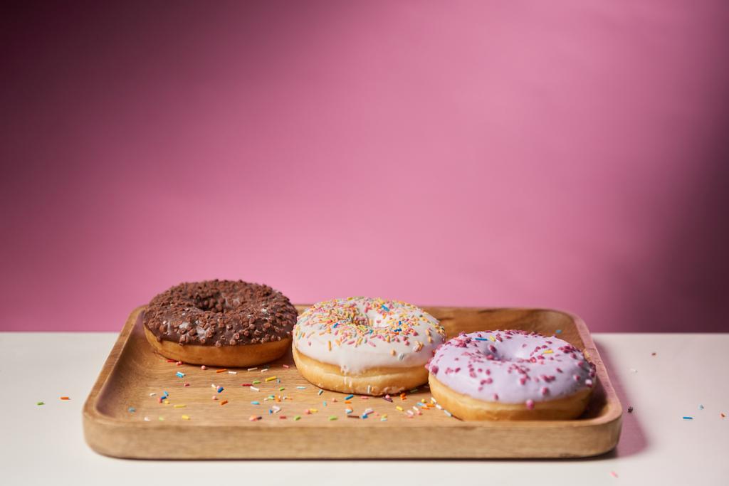 вкусные глазурованные пончики посыпаются на деревянную доску для резки на розовом фоне
  - Фото, изображение