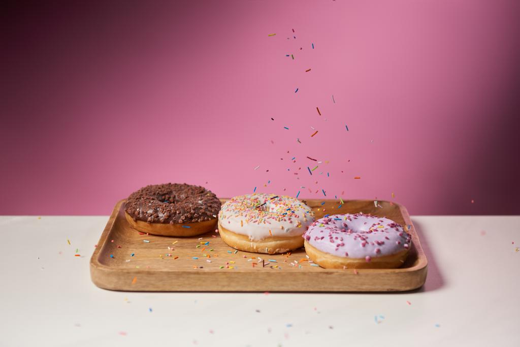 lekkere donuts met vallende hagelslag op houten snijplank op roze achtergrond  - Foto, afbeelding