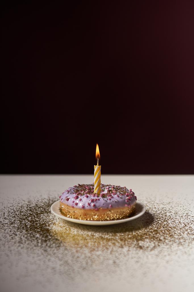 vela ardente no meio de donut envidraçado com brilhos dourados na mesa branca isolada em preto
 - Foto, Imagem