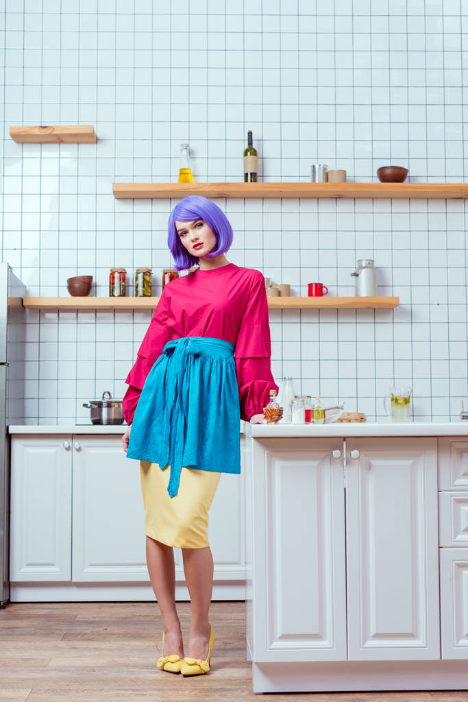 όμορφη νοικοκυρά με μοβ μαλλιά και πολύχρωμα ρούχα, ποζάρει στην κουζίνα  - Φωτογραφία, εικόνα