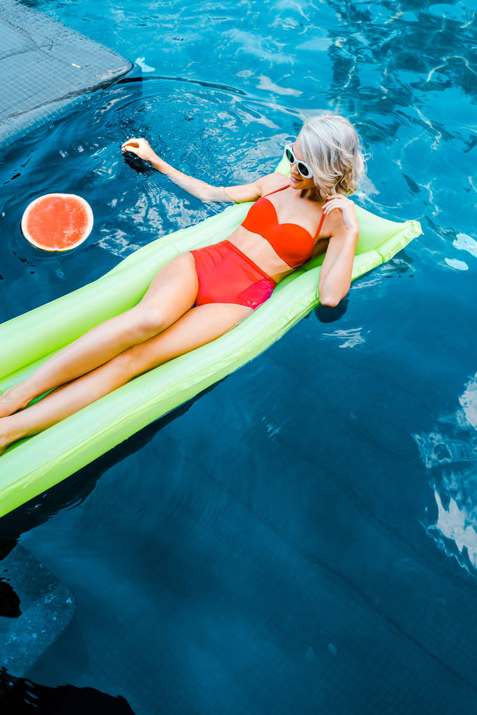 mooi meisje in zwembroek rustend op groene opblaasbare matras in zwembad met watermeloen - Foto, afbeelding