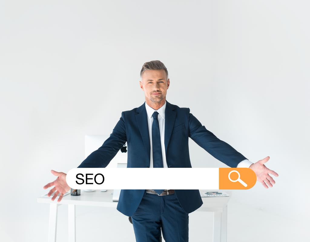 handsome businessman standing with open arms near table with computer isolated on grey with seo in search bar - Photo, Image