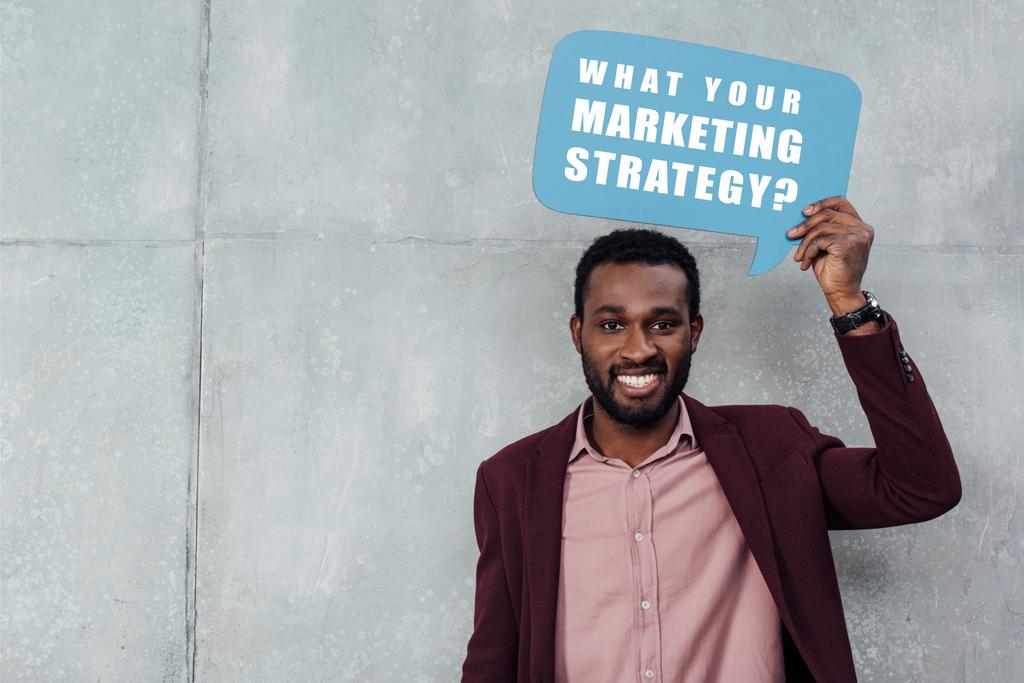 sorridente empresário casual afro-americano olhando para a câmera e segurando bolha de fala com "qual é a sua estratégia de marketing?" lettering em fundo cinza
  - Foto, Imagem