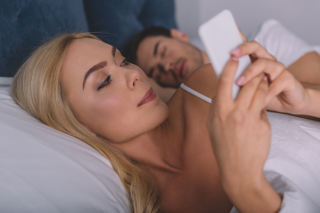 beautiful suspicious woman using smartphone while lying in bed with sleeping husband, secret concept - Photo, Image