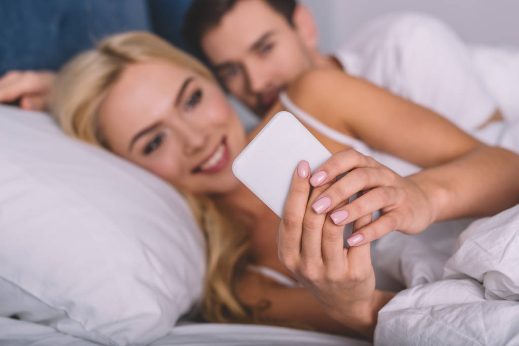 enfoque selectivo del joven que mira a su novia sonriente usando el teléfono inteligente en la cama, concepto de desconfianza
 - Foto, imagen