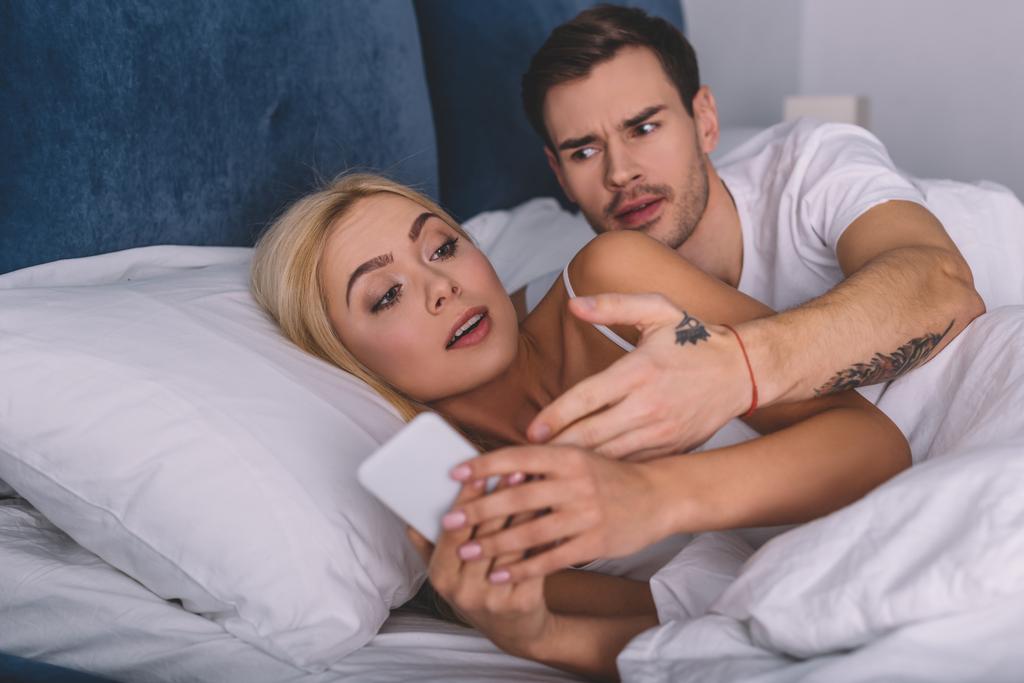 Wütender Mann sieht verdächtige Freundin mit Smartphone im Bett an, misstraut Konzept - Foto, Bild