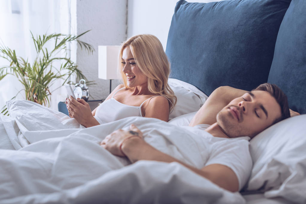 guapo joven hombre durmiendo y hermosa mujer sonriente usando teléfono inteligente en la cama
 - Foto, Imagen