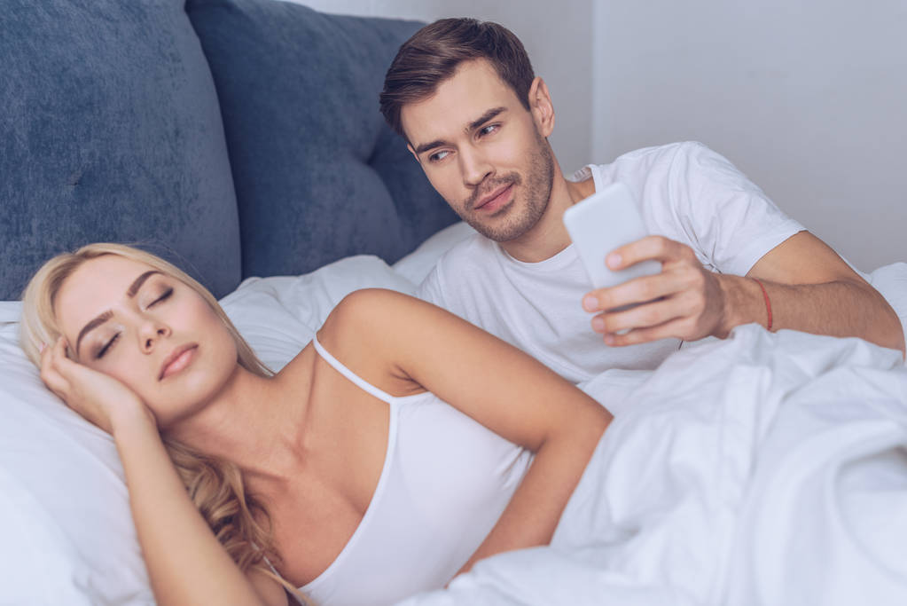 усміхнений молодий чоловік тримає смартфон і дивиться на сплячу дівчину в ліжку, секретна концепція
 - Фото, зображення