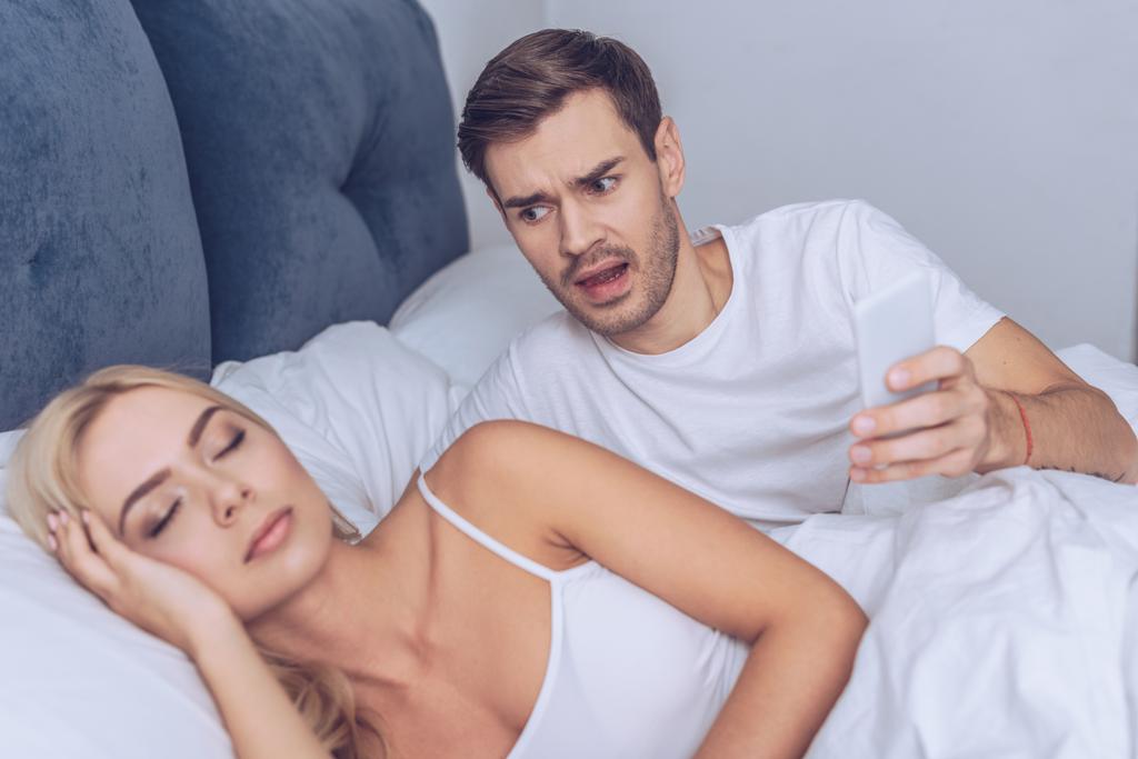 Angry young man houden smartphone en slapende meisje in bed te kijken - Foto, afbeelding