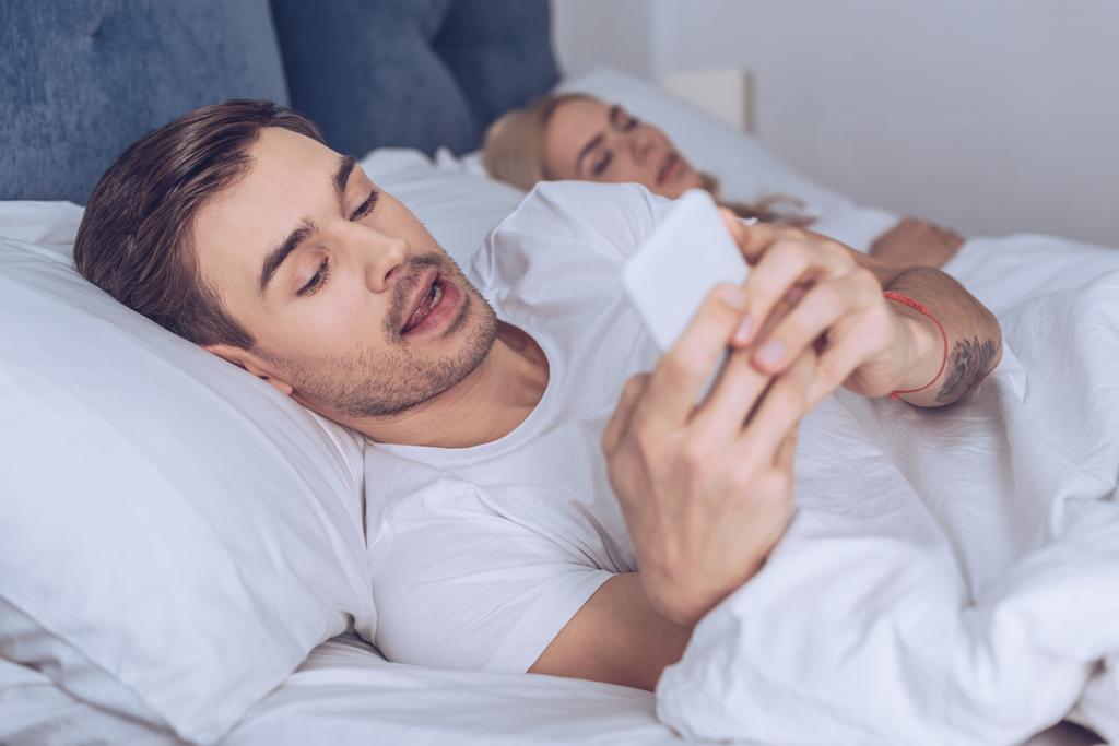 joven sospechoso usando un teléfono inteligente mientras está acostado con su esposa dormida en la cama
  - Foto, Imagen