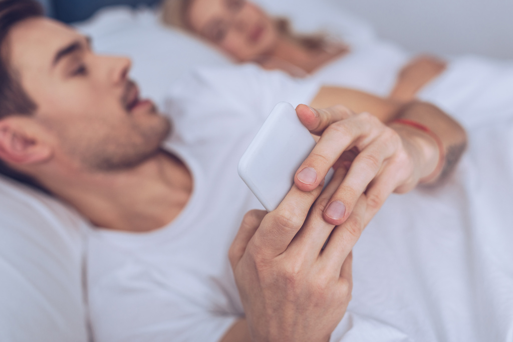 Крупный план молодого человека с помощью смартфона, лежащего со спящей женой в постели
 - Фото, изображение