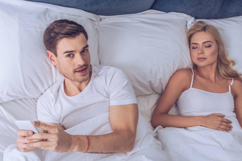 високий кут зору молодого чоловіка за допомогою смартфона і дивиться на камеру, лежачи зі сплячою дружиною в ліжку
 - Фото, зображення