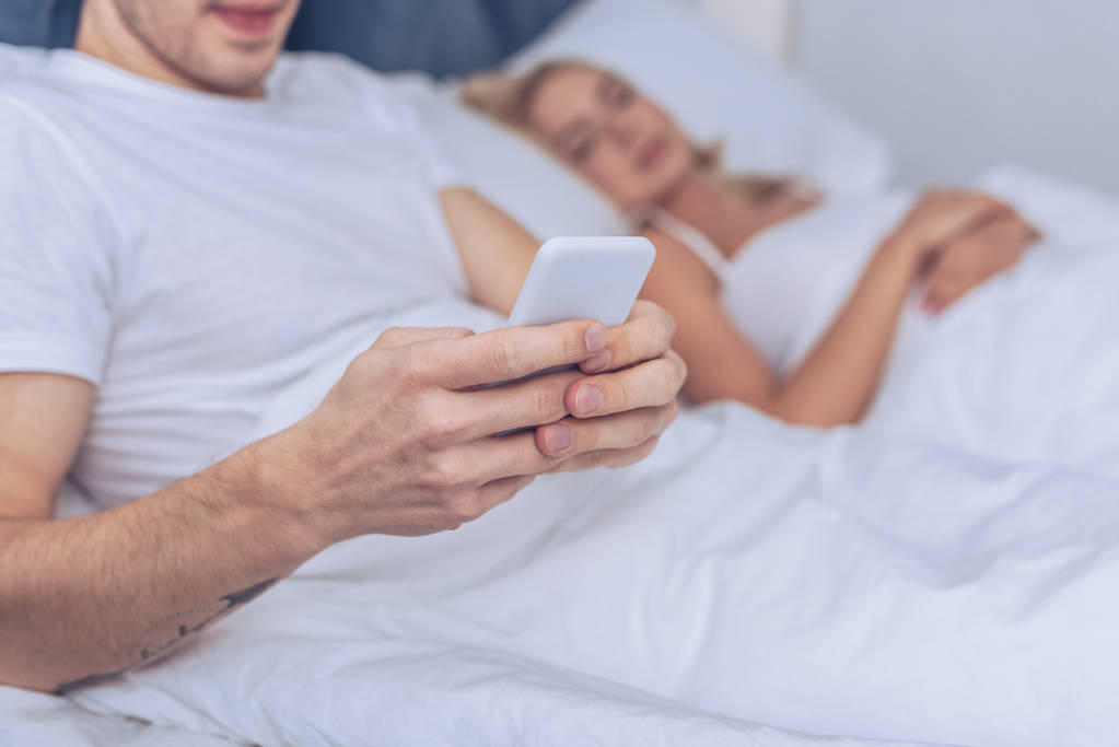 bijgesneden schot van jonge man met behulp van smartphone liggend met slapende vrouw in bed - Foto, afbeelding