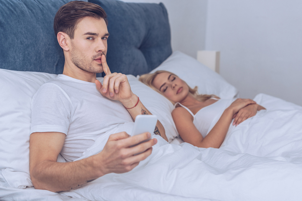 homem com gesto smartphone para o silêncio e olhando para a câmera enquanto deitado com a esposa dormindo na cama
 - Foto, Imagem
