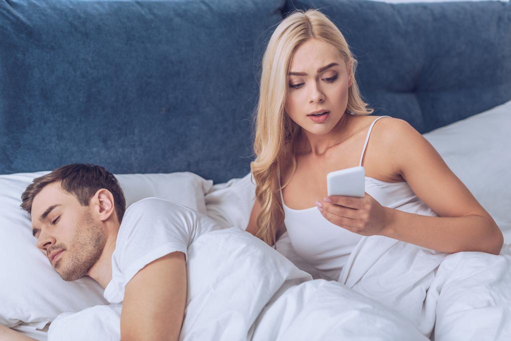 mustasukkainen vaimo käyttää älypuhelinta, kun aviomies nukkuu sängyssä, epäluottamus käsite
 - Valokuva, kuva