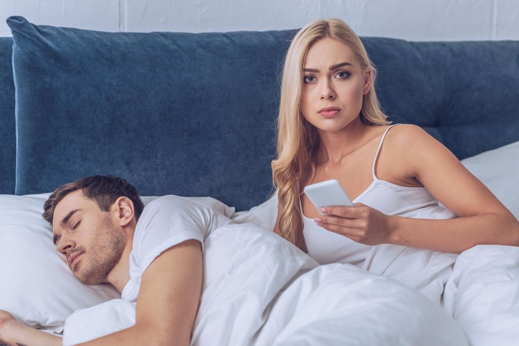 ongelukkige jonge vrouw houden van smartphone en kijken naar de camera terwijl vriendje slapen in bed, wantrouwen concept - Foto, afbeelding