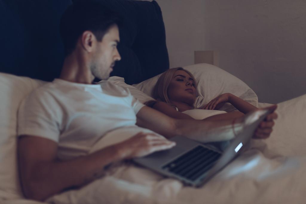 epäilyttävä nuori mies käyttää kannettavaa tietokonetta ja katselee tyttöystävä nukkuu sängyssä yöllä
   - Valokuva, kuva