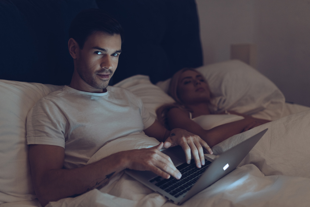 epäilyttävä nuori mies käyttää kannettavaa tietokonetta ja katselee kameraa, kun vaimo nukkuu sängyssä yöllä
  - Valokuva, kuva