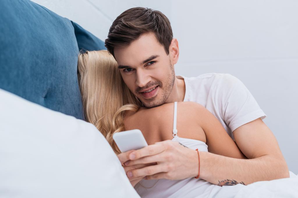 young man using smartphone and smiling at camera while lying with girlfriend in bed - Photo, Image