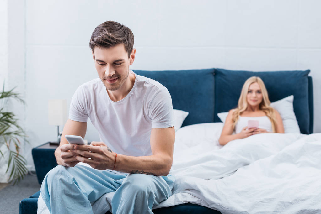 Nuori nainen makaa sängyssä älypuhelimen ja miehensä kanssa älypuhelimen avulla etualalla makuuhuoneessa
 - Valokuva, kuva