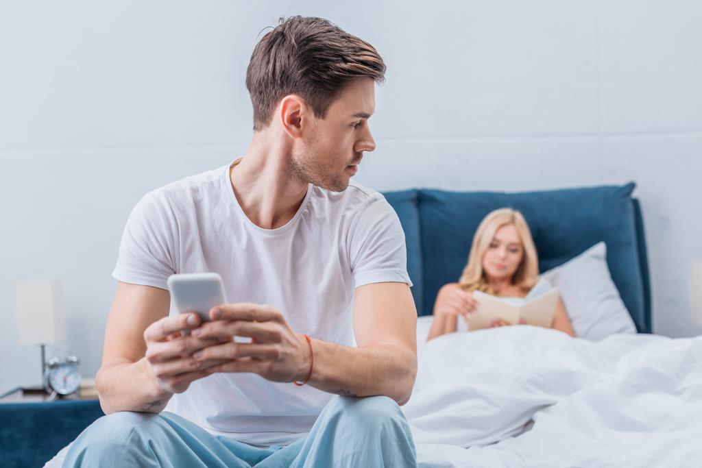 komea nuori mies pyjamassa älypuhelimella ja katsomalla vaimoa lukemassa kirjaa sängyssä, epäluottamus käsite
 - Valokuva, kuva