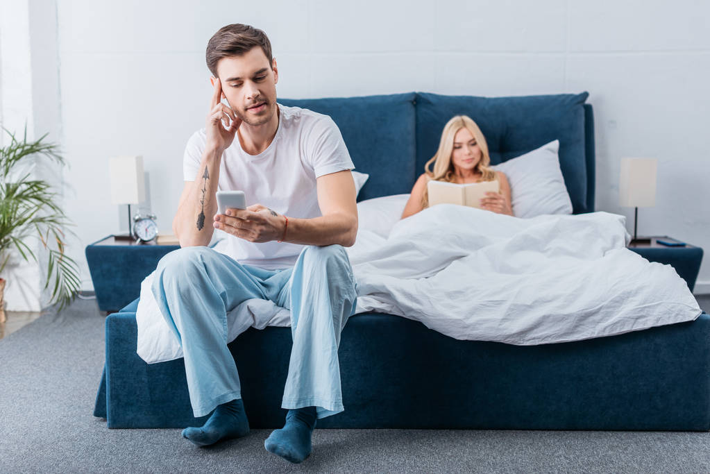 pohledný mladý muž sedí na posteli a pomocí smartphone zároveň přítelkyně čtení knih v posteli - Fotografie, Obrázek