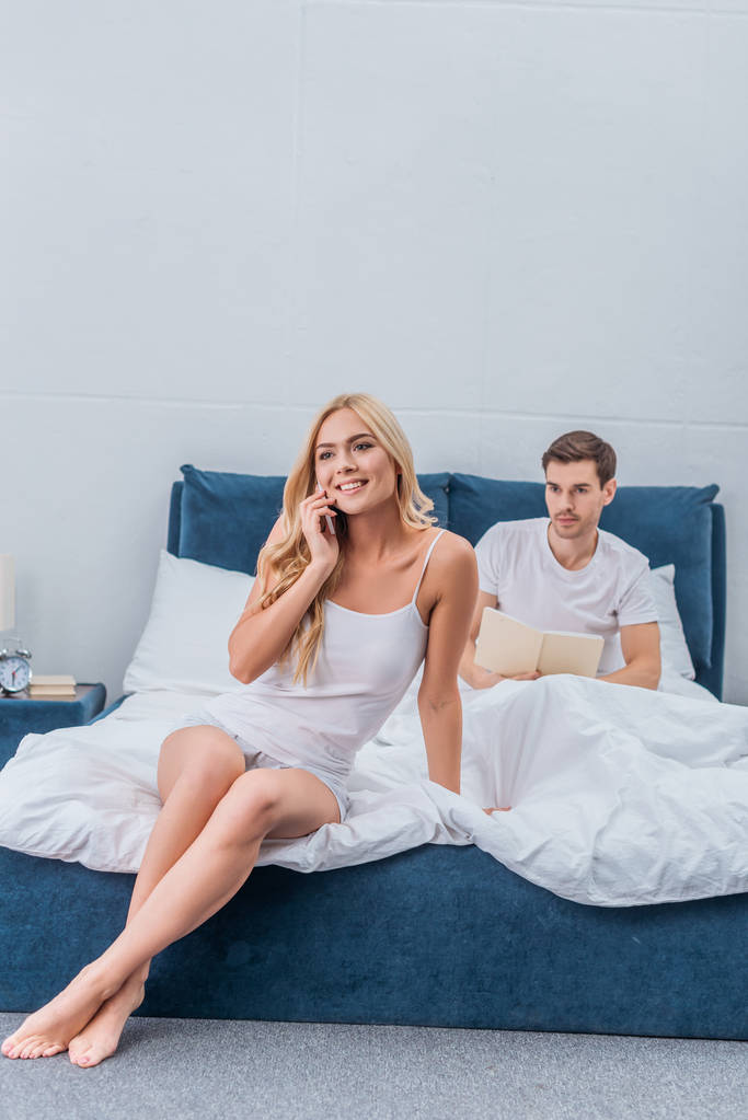 hermosa mujer sonriente hablando por teléfono inteligente mientras novio celoso leyendo libro detrás en la cama
   - Foto, Imagen