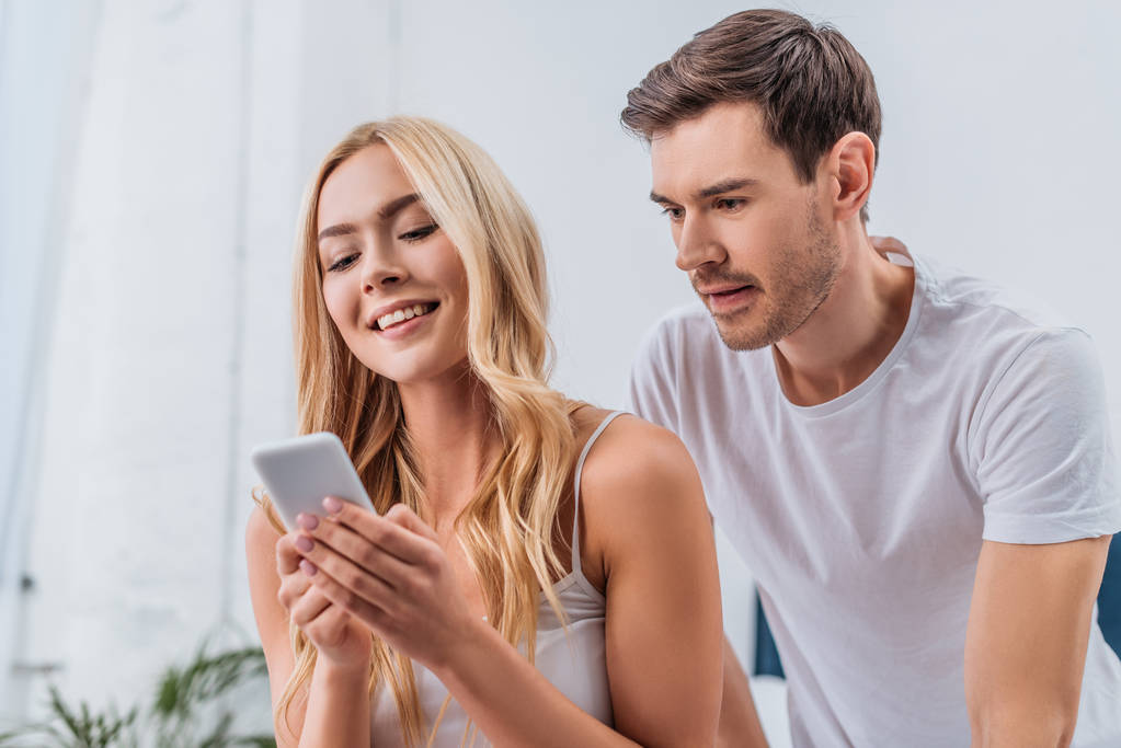 jealous young man looking at beautiful smiling girlfriend using smartphone in bedroom  - Photo, Image