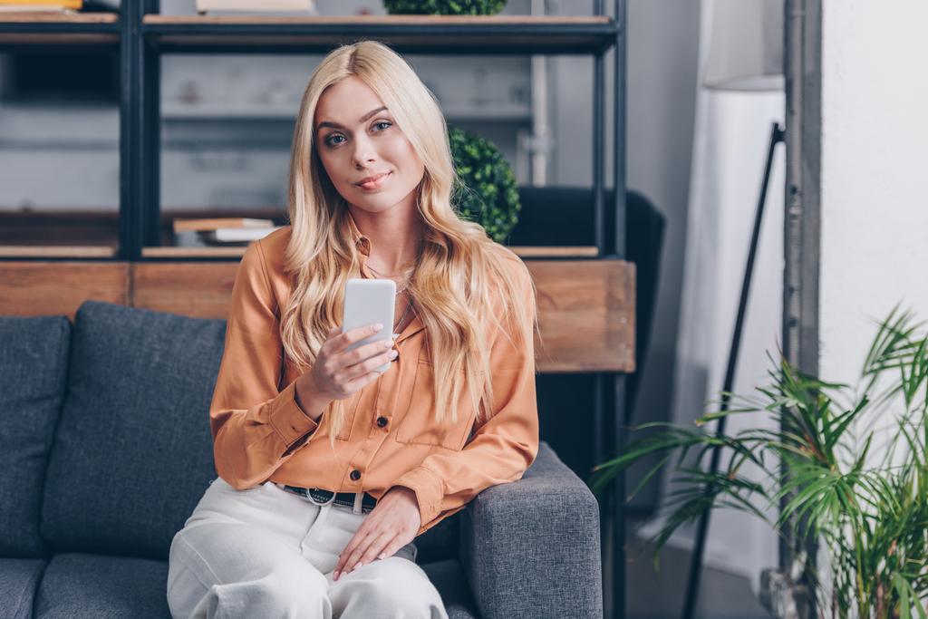 mooie blonde vrouw met behulp van smartphone en glimlachend in de camera terwijl zittend op de Bank thuis - Foto, afbeelding