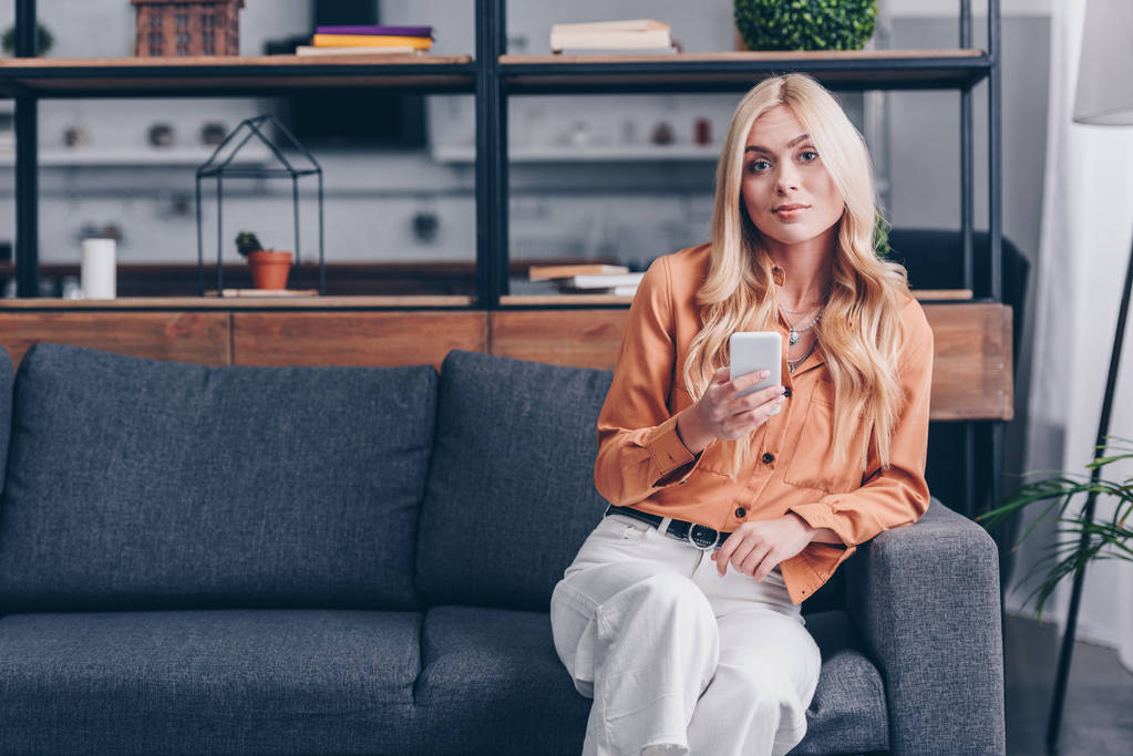 attraktive blonde Mädchen mit Smartphone und lächelt in die Kamera, während sie zu Hause auf der Couch sitzt - Foto, Bild