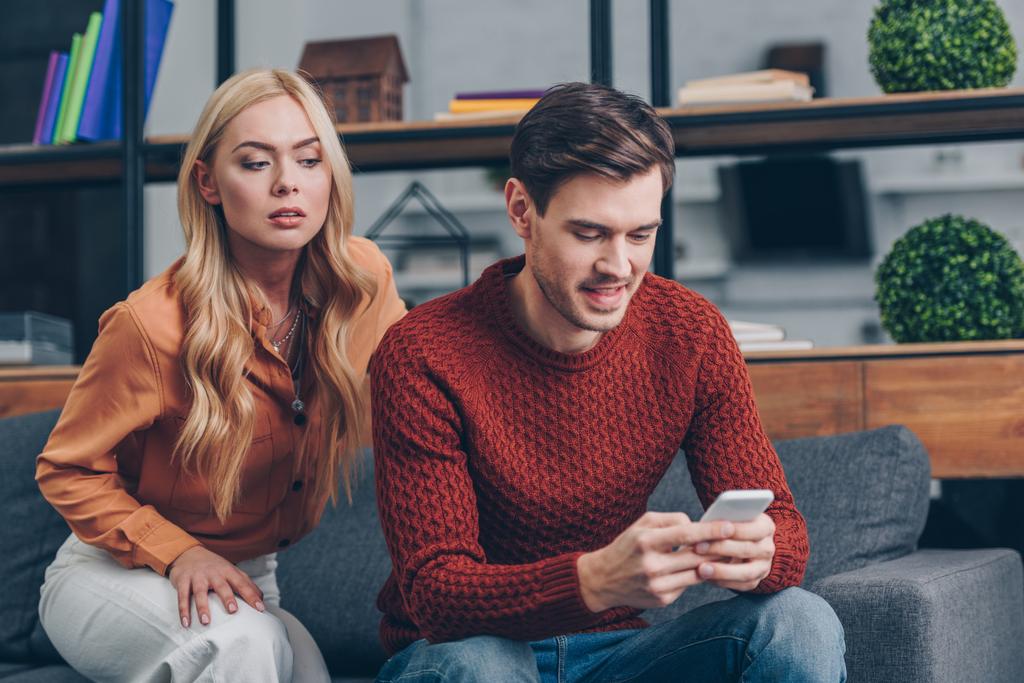 jealous young woman looking at smiling husband using smartphone on couch, distrust concept - Photo, Image