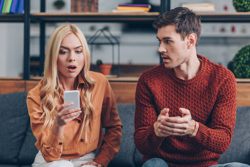 emotional young man looking at shocked girlfriend using smartphone on couch, jealousy concept - Photo, Image
