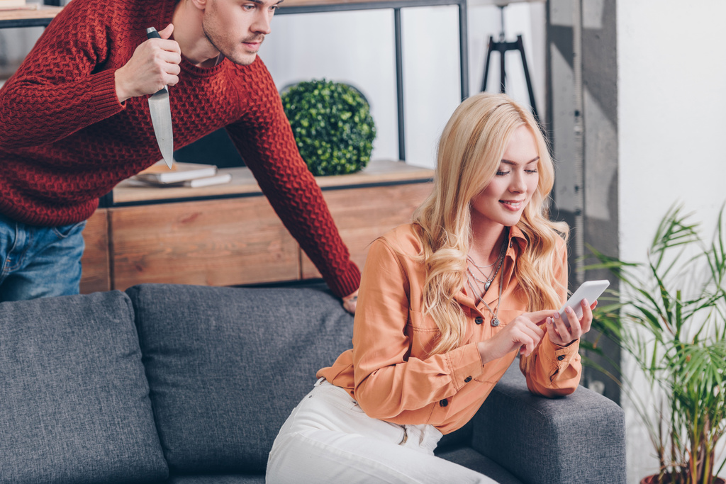cropped shot of aggressive man with knife looking at smiling wife using smartphone on couch, jealousy concept  - Photo, Image