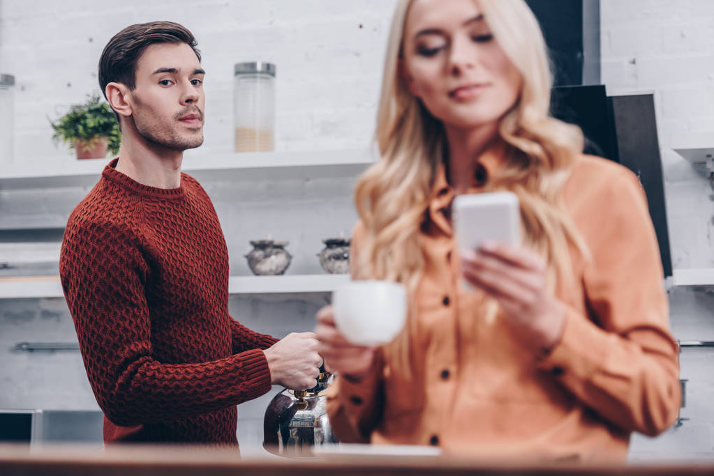Selektywny fokus zazdrosny człowiek ogląda uśmiechnięta dziewczyna gospodarstwa Puchar i przy użyciu smartfona w kuchni - Zdjęcie, obraz
