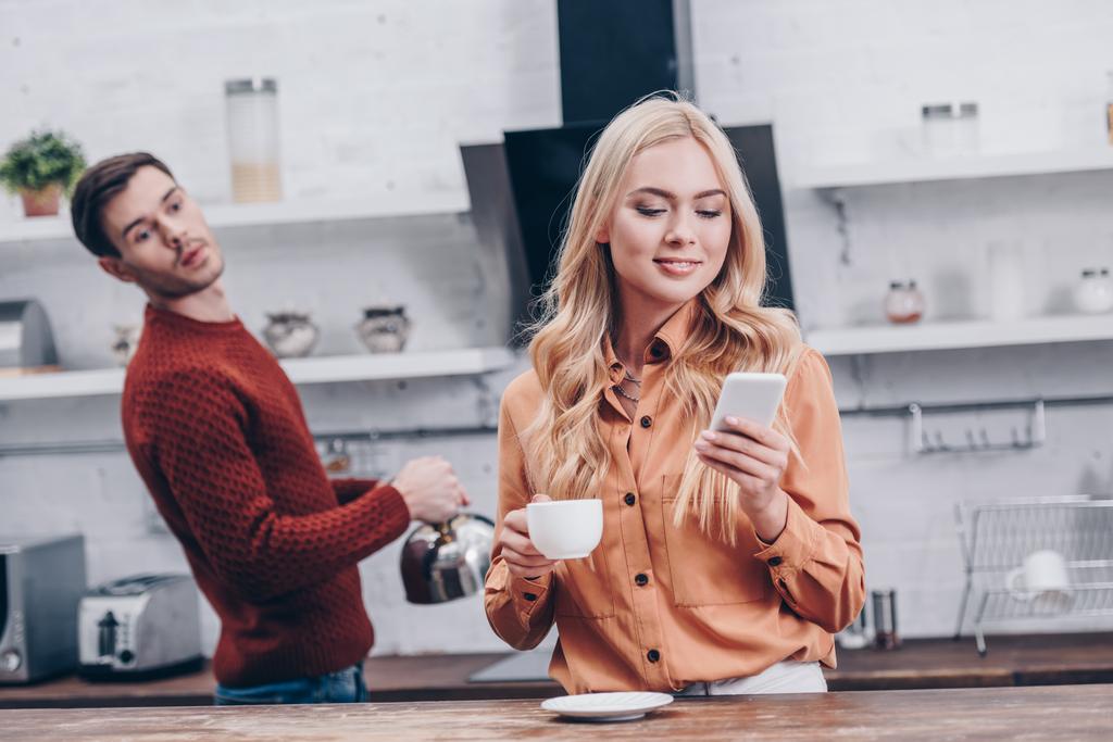 kıskanç genç adam kız arkadaşı holding Kupası gülümseyen ve mutfakta smartphone kullanarak bakarak - Fotoğraf, Görsel