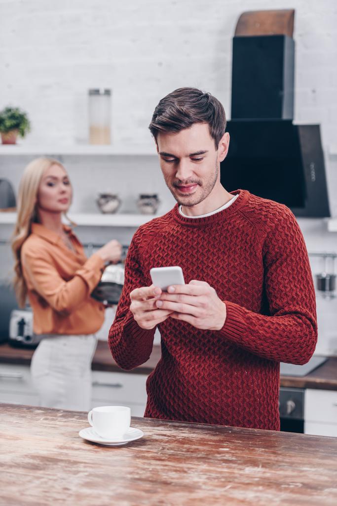ревнивая женщина смотрит на улыбающегося мужа с помощью смартфона на кухне, концепция недоверия
 - Фото, изображение