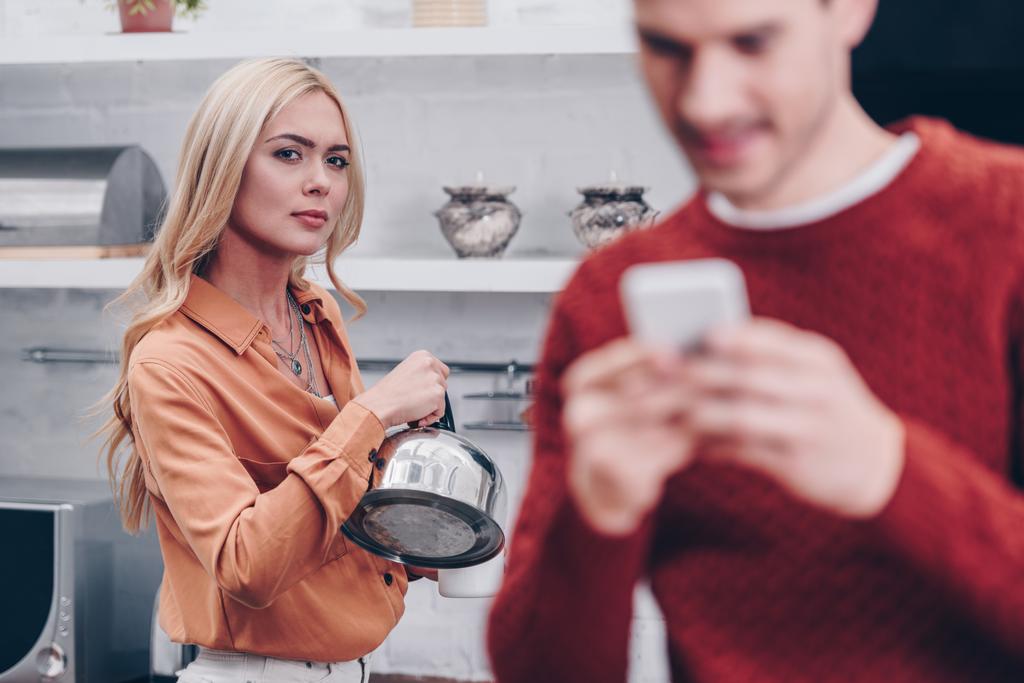 Selektivní fokus žárlivý mladá žena s konvicí na manžela pomocí smartphone v kuchyni  - Fotografie, Obrázek